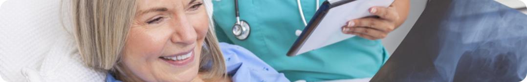 A woman looks at her xray after surgery. She is smiling. A nurse stands by with a clipboard in hand.